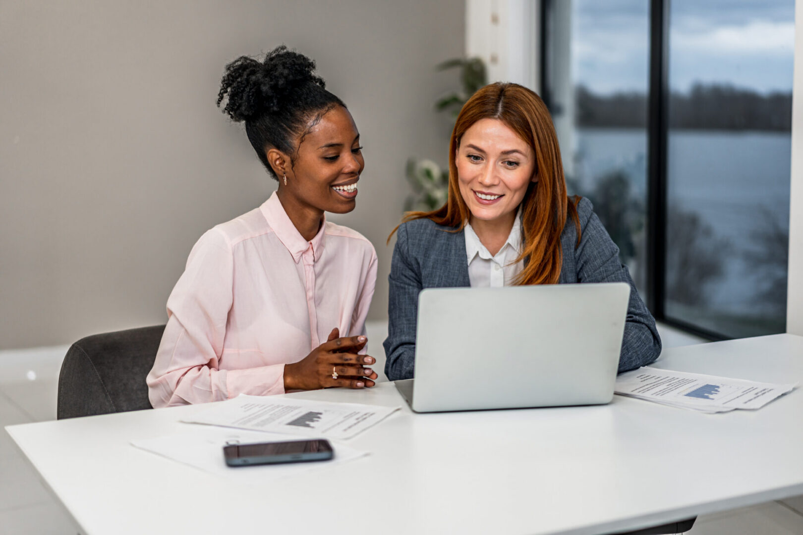 Two smiling women are working together in a corporate setting, reviewing information on a laptop, showing teamwork and partnership